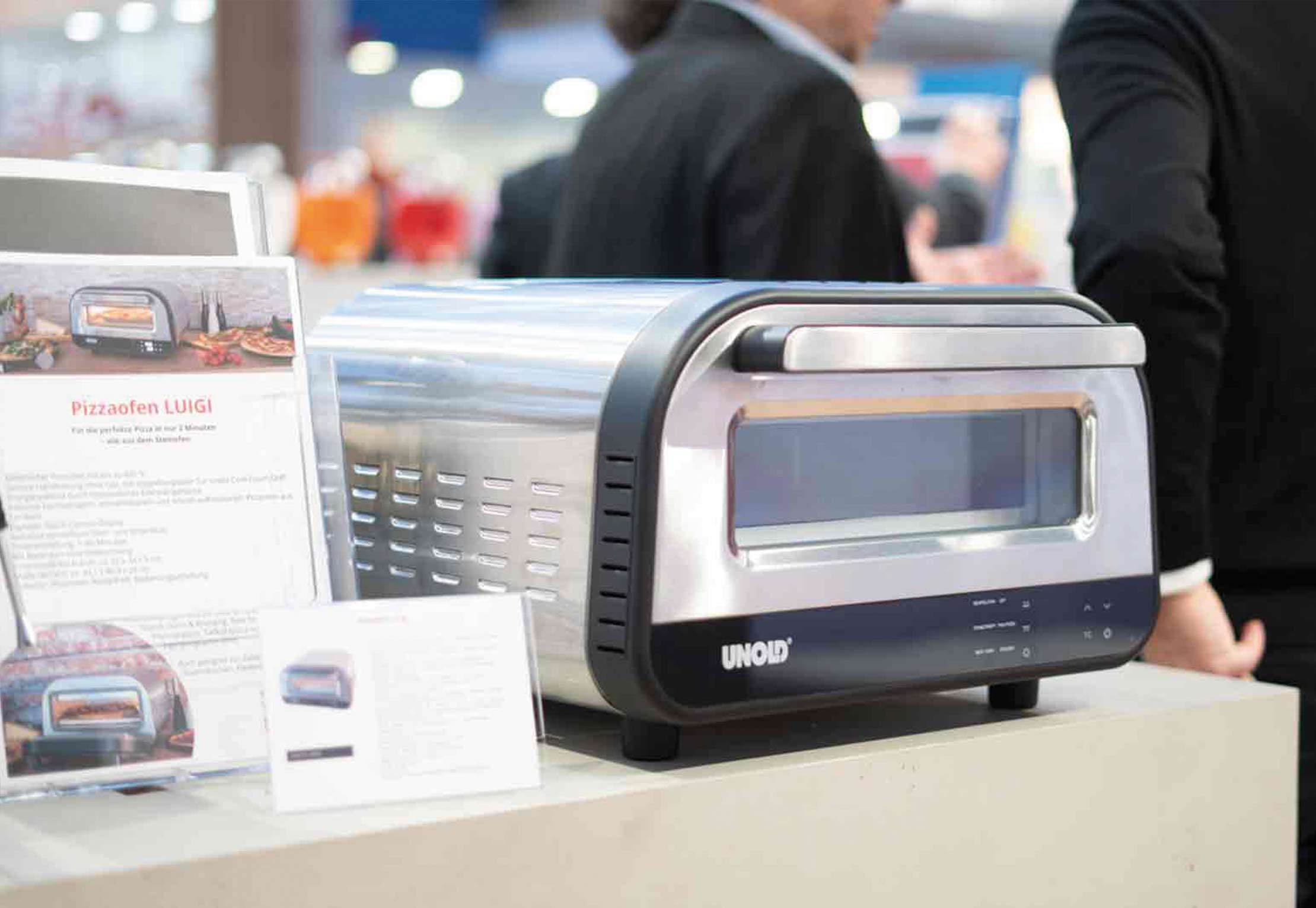 Ein kompakter Edelstahl-Pizzaofen von UNOLD steht auf einem Tresen bei einer Veranstaltung. Daneben liegt ein Informationsblatt. Im unscharfen Hintergrund sind Personen in Business-Kleidung zu sehen.