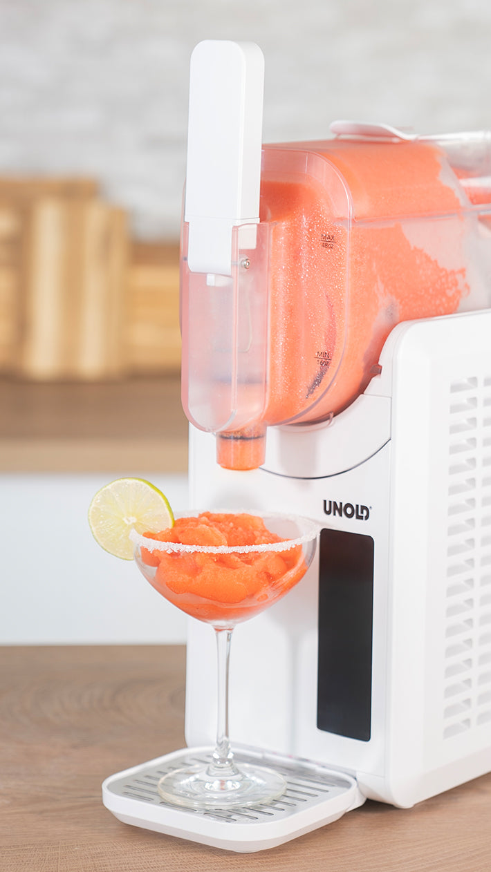 Eine weiße Slushie-Maschine schüttet Orangensaft in ein Cocktailglas, das mit einer Limettenscheibe und einem gezuckerten Rand garniert ist. Die Maschine steht auf einer Küchentheke mit Holzkanistern im Hintergrund.