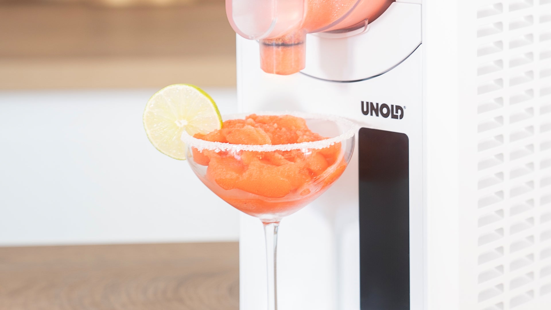 Ein Margarita-Glas mit einem gesalzenen Rand und einer Limettenscheibe wird mit einem gefrorenen Orangengetränk gefüllt, das aus einem weißen UNOLD-Automaten auf einem Küchentresen ausgegeben wird.