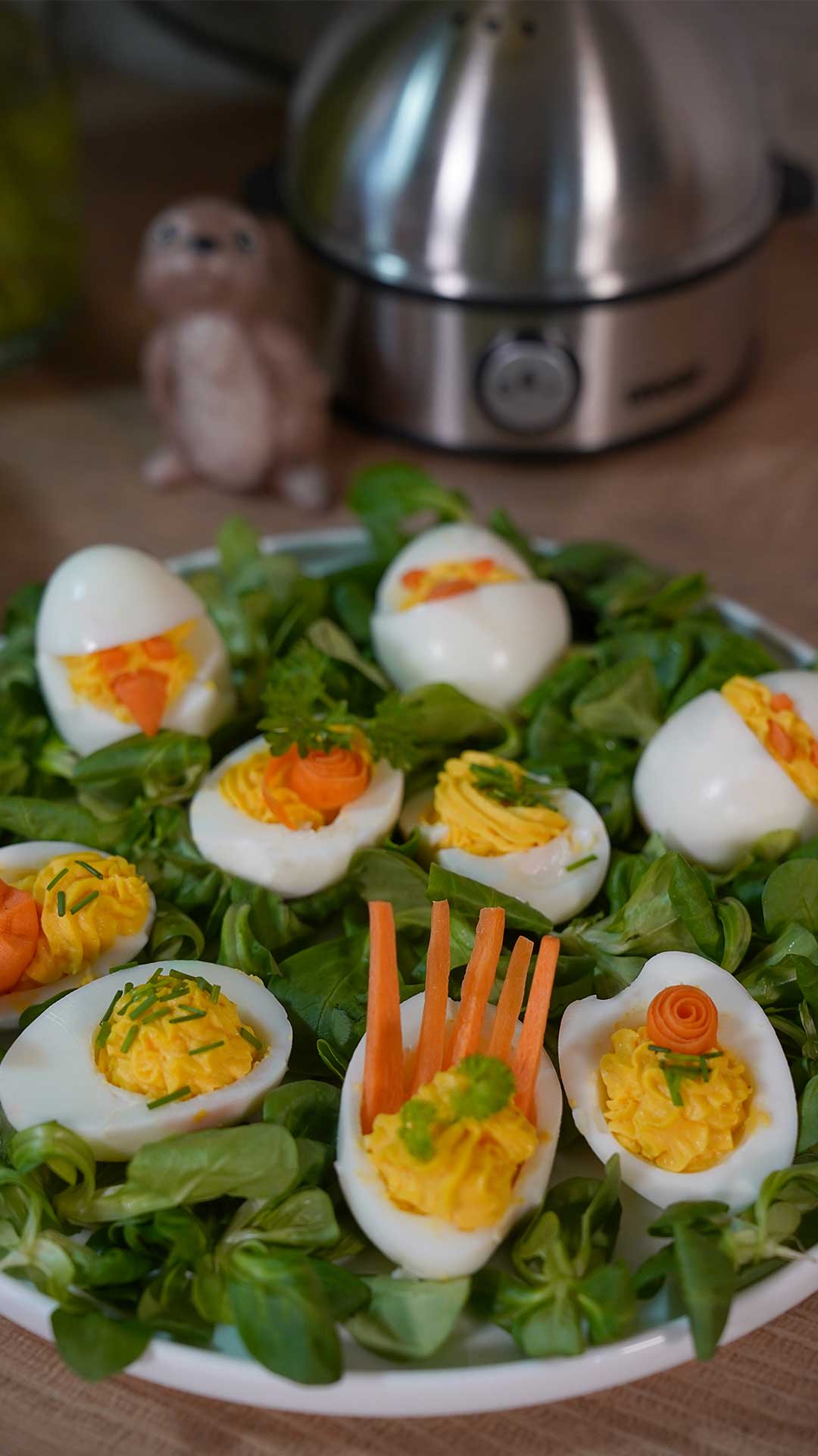 Ein Teller gefüllter Eier, dekoriert mit Kräutern und Karottenmotiven, steht auf einem Bett aus Grünzeug. Im Hintergrund stehen ein Eierkocher aus Metall und eine kleine Figur auf einer Küchentheke.