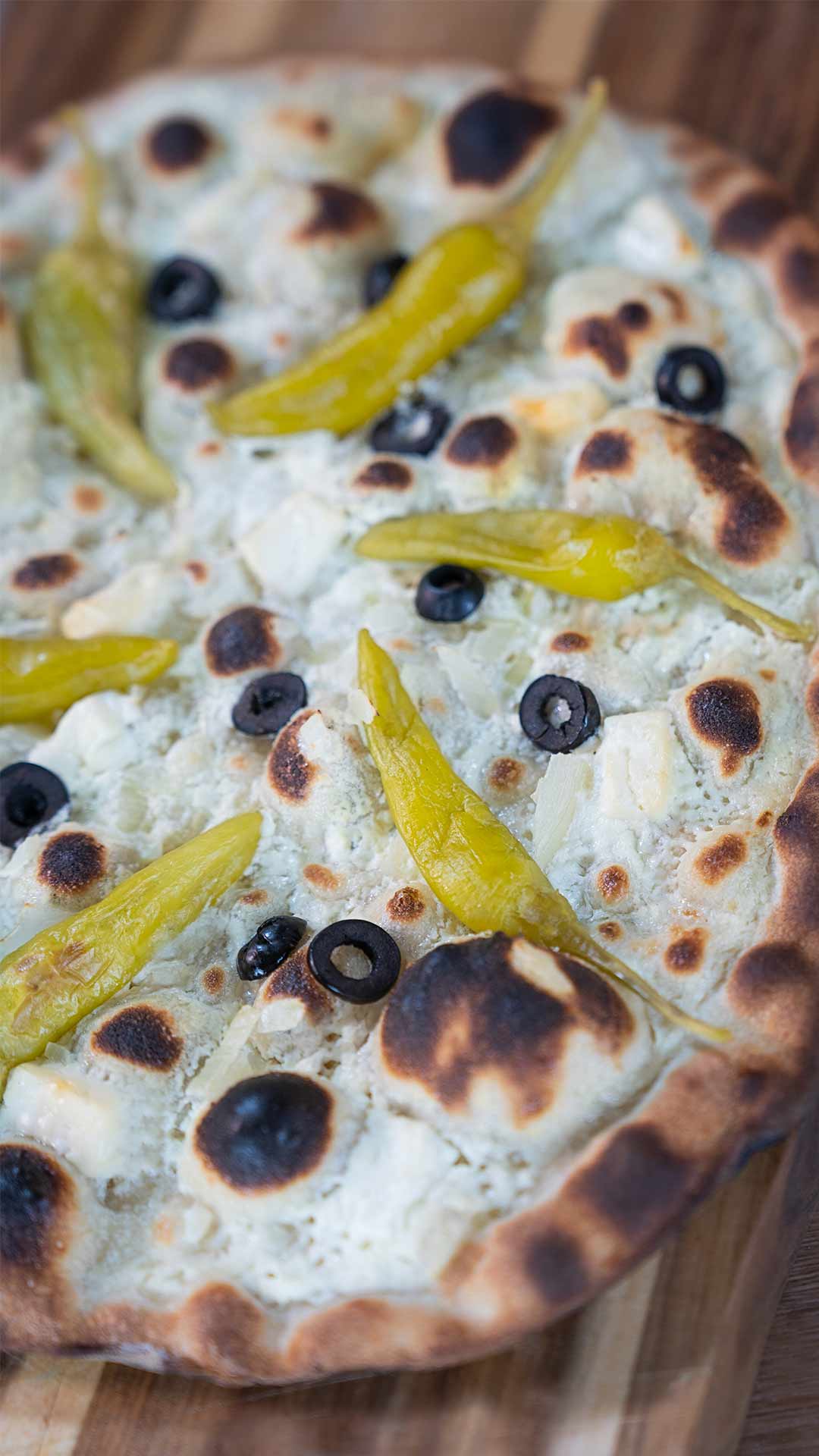 Eine Nahaufnahme einer gebackenen Pizza mit dünnem Boden, belegt mit geschmolzenem Käse, schwarzen Olivenscheiben und ganzen gelben Peperoncini-Paprika auf einer Holzoberfläche.