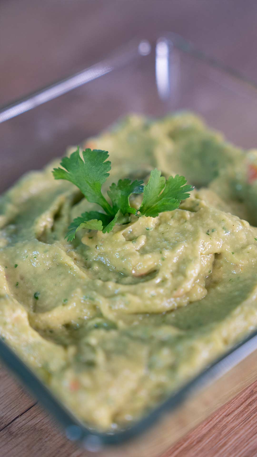Eine Nahaufnahme von cremiger Guacamole, garniert mit frischem Koriander, serviert in einer durchsichtigen Glasschale auf einer Holzoberfläche.