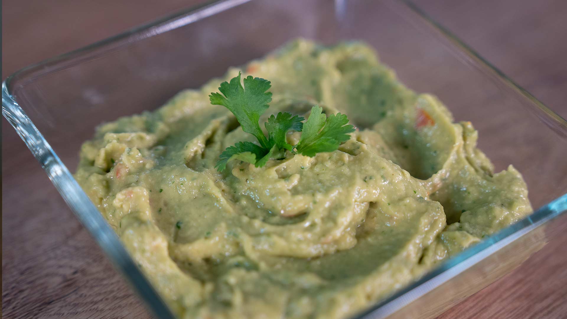 Auf einer Holzoberfläche steht eine durchsichtige Glasschale mit cremiger Guacamole und einem Zweig frischem Koriander.