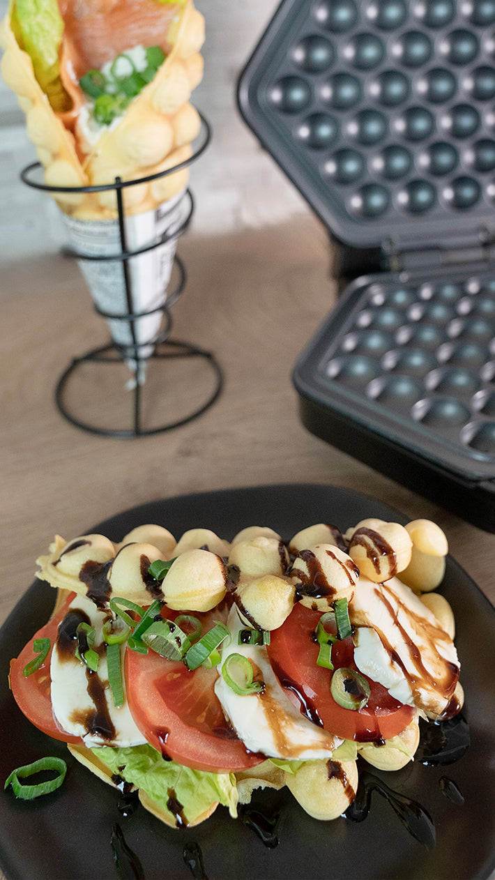 Eine Bubble-Waffel mit Tomatenscheiben, Mozzarella, Salat, Frühlingszwiebeln und dunkler Sauce auf einem schwarzen Teller. Im Hintergrund sind eine Bubble-Waffel mit Lachs und Gemüse sowie ein Bubble-Waffel-Eis zu sehen.