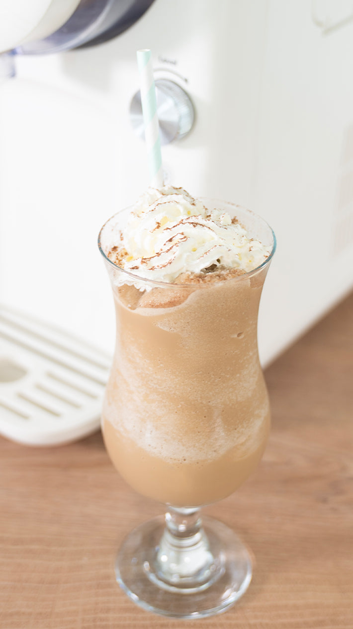 Ein Glas Eiskaffee mit Schlagsahne und einer Prise Kakao, serviert mit einem blau gestreiften Strohhalm, steht auf einer Holzplatte vor einer Kaffeemaschine.