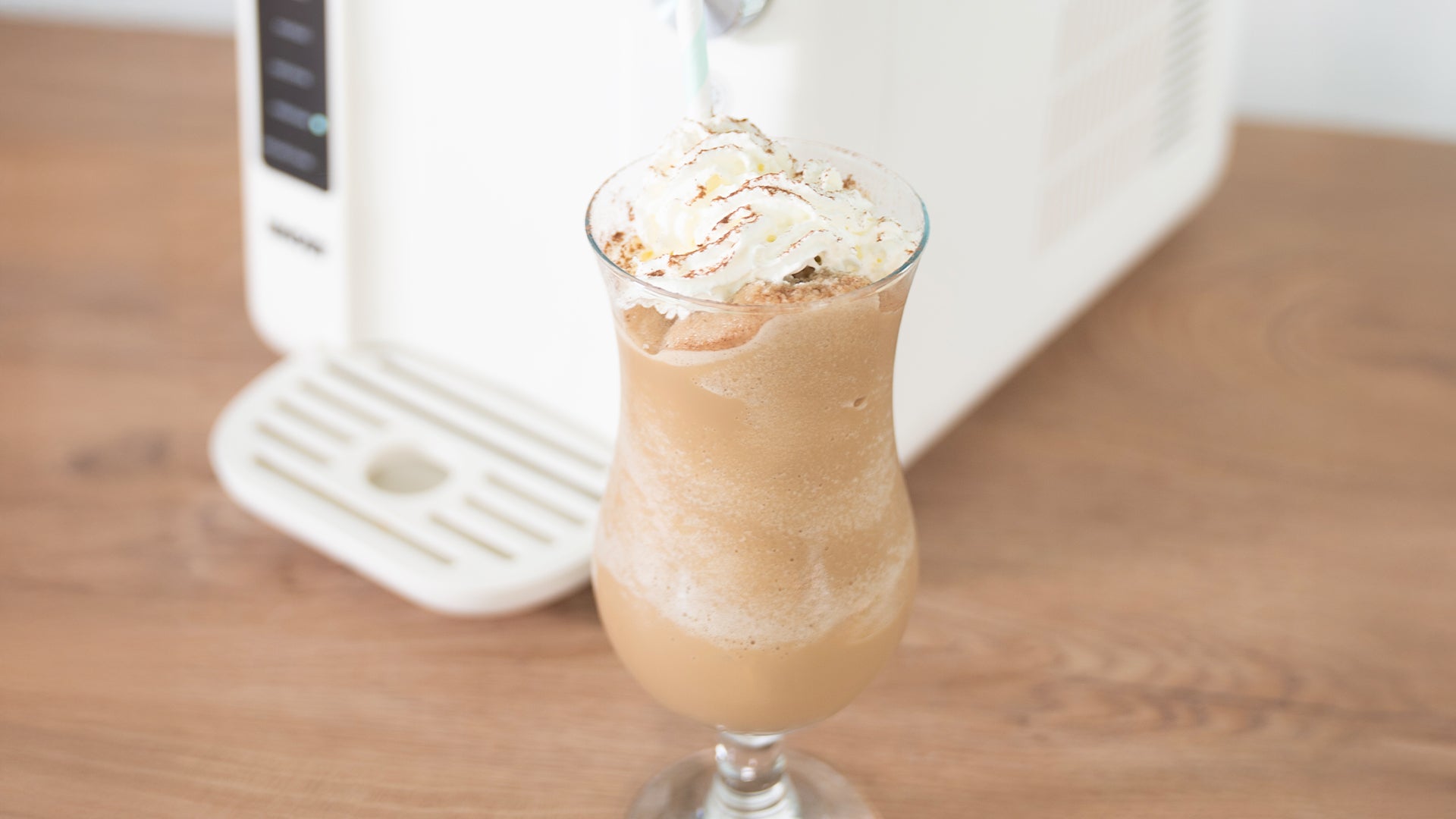 Ein hohes Glas Eiskaffee mit Schlagsahne steht auf einer Holzplatte neben einem weißen Gerät.