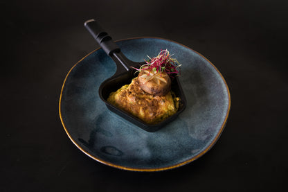 Das Unold Raclette Finesse Basic inklusive Raclette Buch wird in einer kleinen schwarzen Pfanne auf einem blauen Teller präsentiert und präsentiert vor einem dunklen Hintergrund eine goldbraun gebackene Spezialität, belegt mit Pilzen und leuchtenden Microgreens.