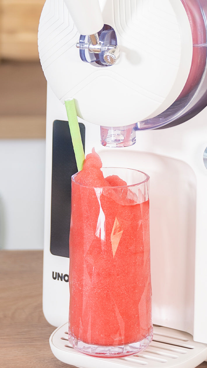 Ein mit leuchtend rotem Slushie gefülltes Glas steht unter einem weißen Slushie-Automaten, mit einem grünen Strohhalm im Getränk. Die Maschine steht auf einer Holztheke.