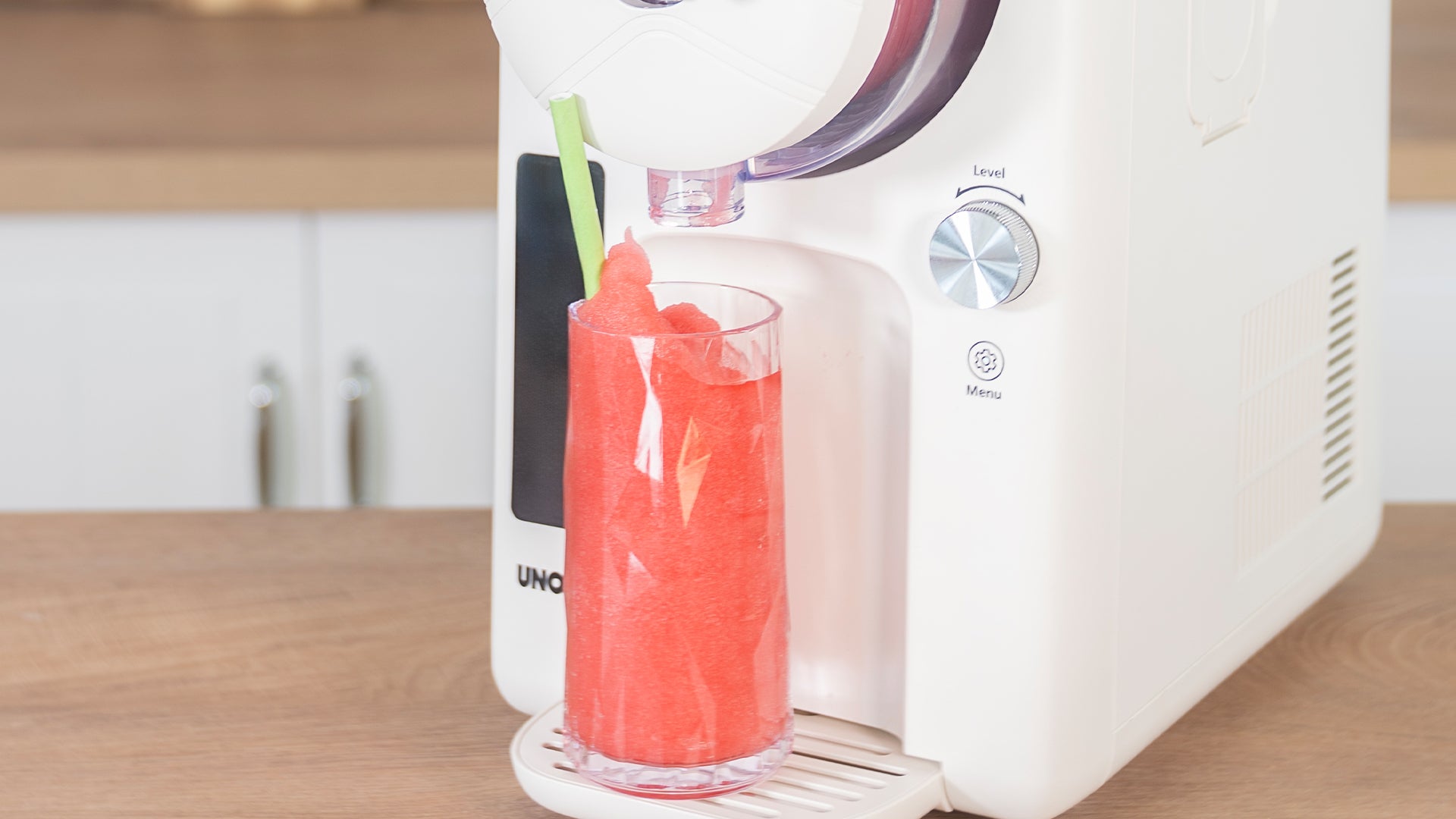 Eine Tischmaschine gibt leuchtend roten Slushy in ein klares Glas mit grünem Strohhalm aus, das auf einer Holzfläche in einer modernen Küche steht.