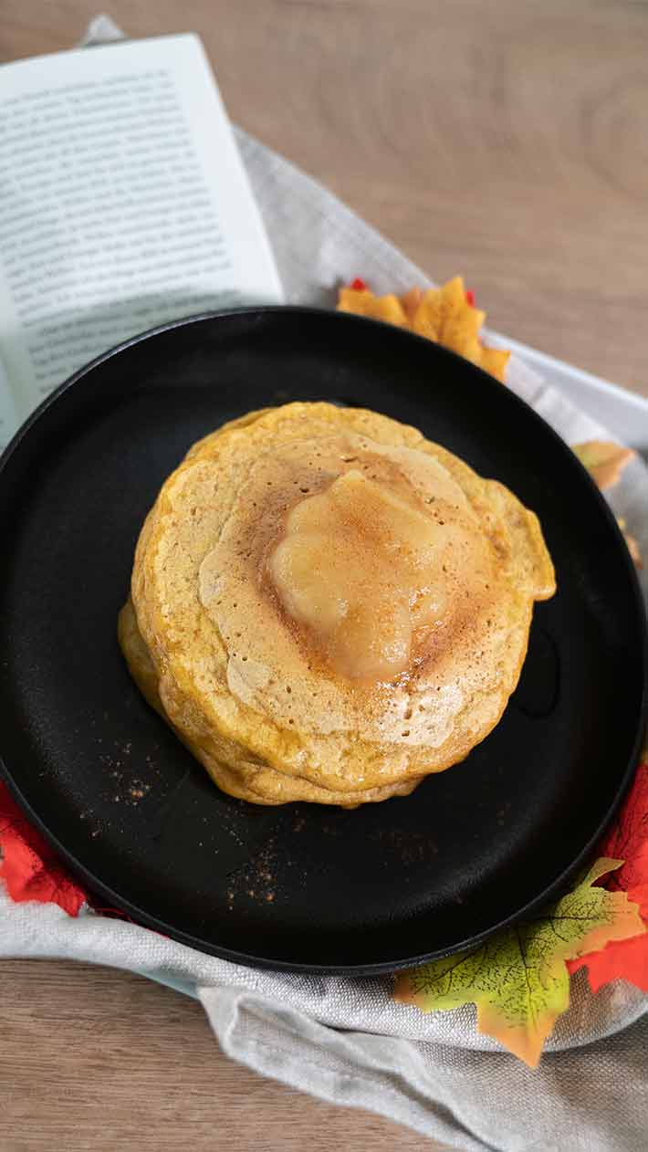 Ein Stapel Pfannkuchen mit einem Klecks Butter liegt auf einem schwarzen Teller. Ein offenes Buch und Herbstblätter liegen neben dem Teller auf einer hellen Oberfläche.