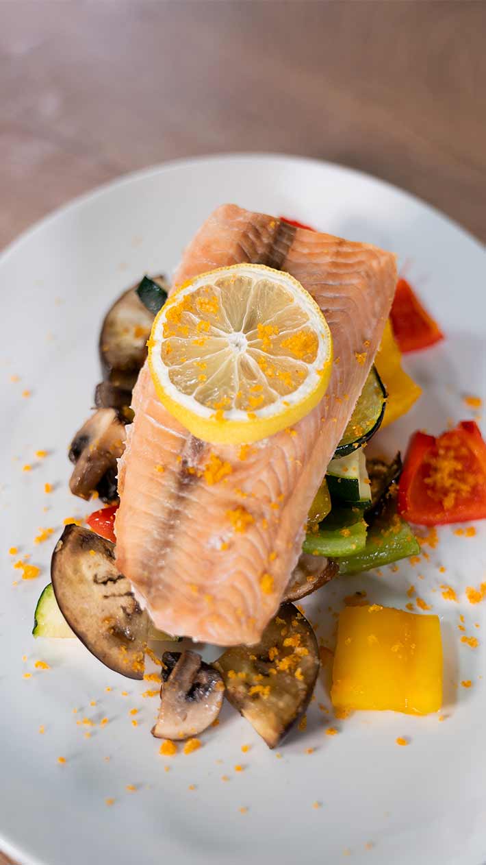 Ein Filet von gekochtem Lachs mit einer Zitronenscheibe, serviert auf einem Bett aus verschiedenen gerösteten Gemüsesorten, darunter Pilze, Zucchini und Paprika, auf einem weißen Teller.