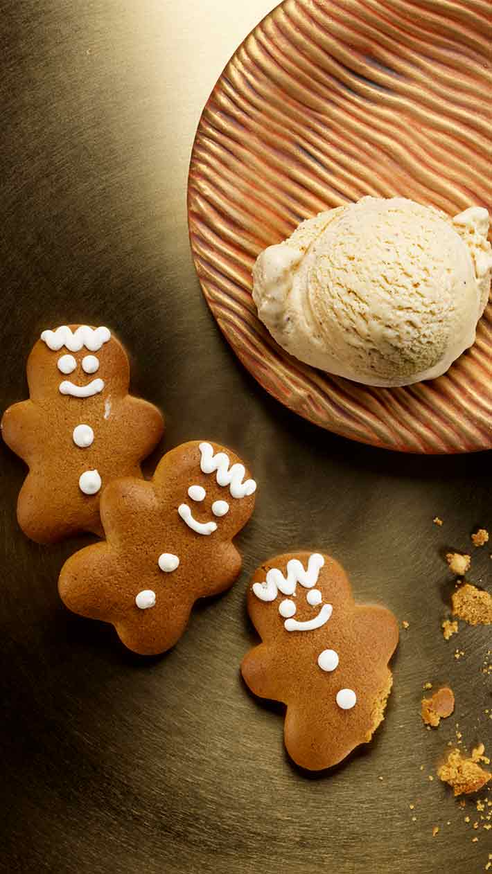 Drei Lebkuchen mit Gesichtern und Knöpfen aus weißem Zuckerguss liegen neben einem Teller mit einer Kugel Vanilleeis. Die Kekse liegen auf einer goldenen Oberfläche, daneben liegen einige Krümel.
