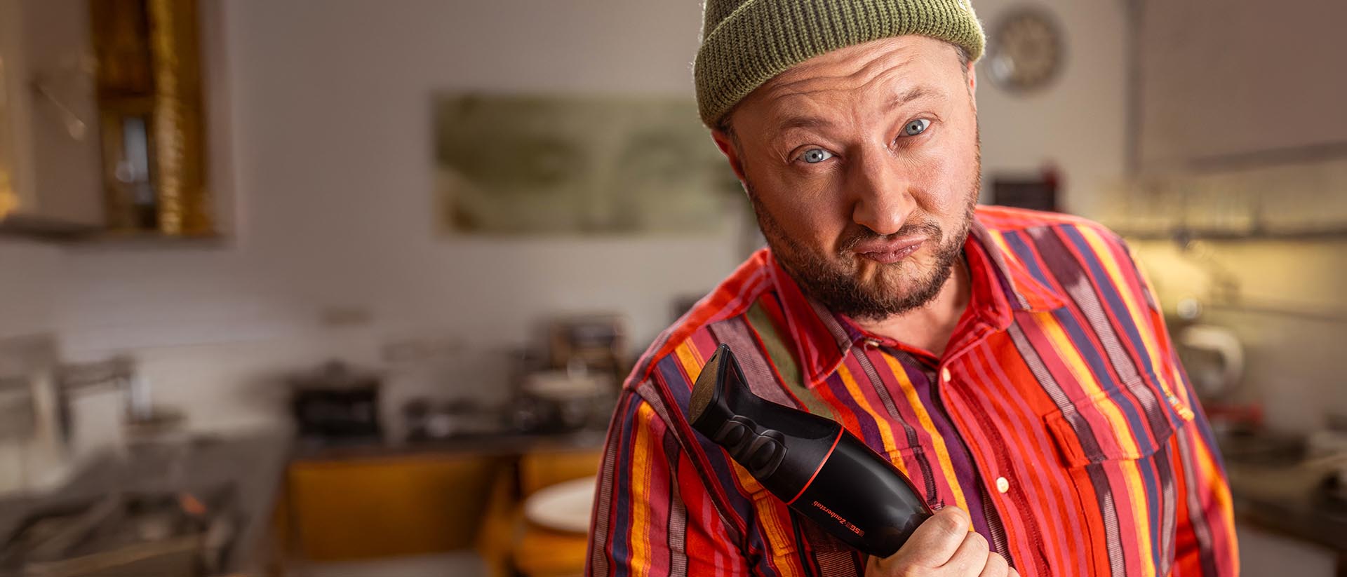 Ein Mann mit Bart, einer grünen Mütze und einem bunt gestreiften Hemd hält einen elektrischen Haarschneider in der Hand und schneidet in einer Küchenumgebung ein verspieltes Gesicht.