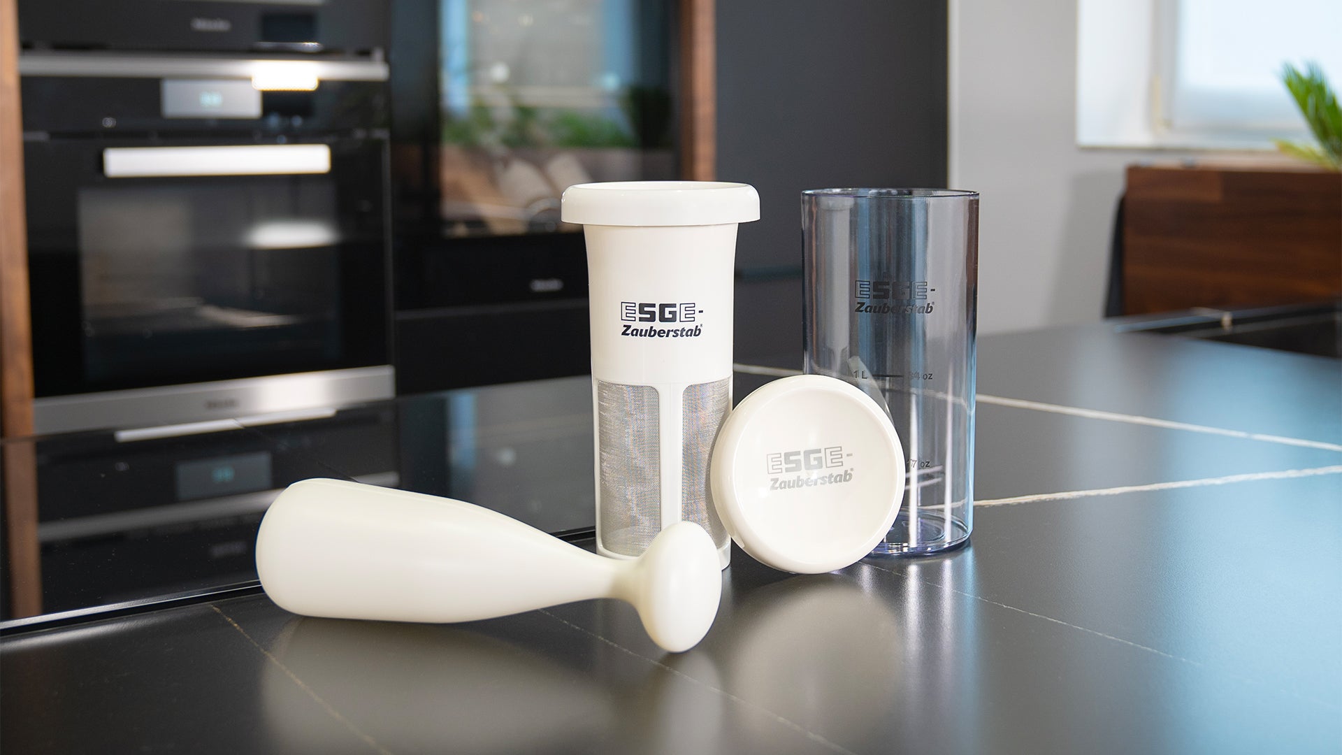 Drei Küchenmixer-Zubehörteile, darunter ein weißer Mixbecher mit Filter, ein durchsichtiger Messbecher und ein weißer Stampfer, werden auf einer modernen schwarzen Arbeitsplatte in einer Küchenumgebung präsentiert.