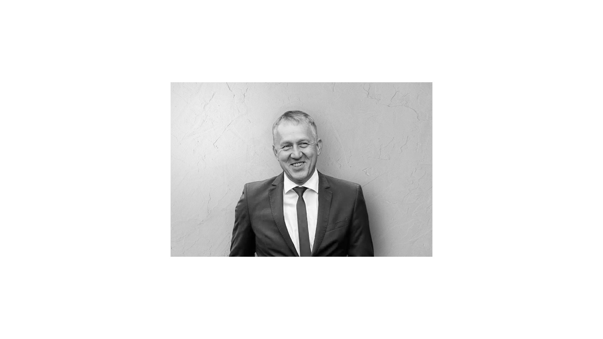Ein Mann in Anzug und Krawatte steht lächelnd vor einer strukturierten Wand. Das Bild ist schwarz-weiß.