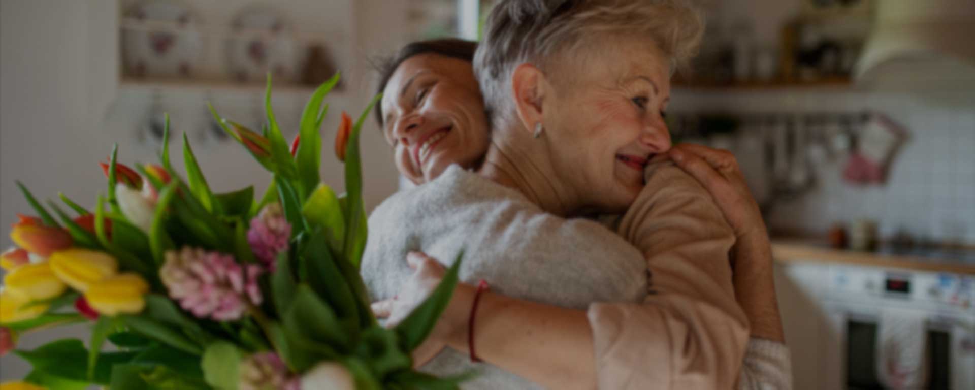 Zwei Frauen umarmen sich und lächeln sich herzlich in einer Küche an. Eine hält einen bunten Tulpenstrauß in der Hand. Der Hintergrund ist sanft verschwommen und schafft so eine gemütliche, fröhliche Atmosphäre.