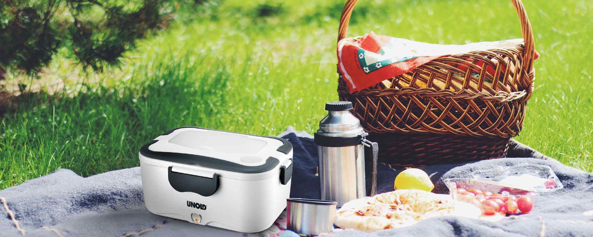 Ein Picknick-Setup auf Gras mit einer Decke, einem Weidenkorb, einer Thermoskanne, einer Tasse, Obst, Trauben, Pizza und einem tragbaren weißen UNOLD-Speisenwärmer im Vordergrund.