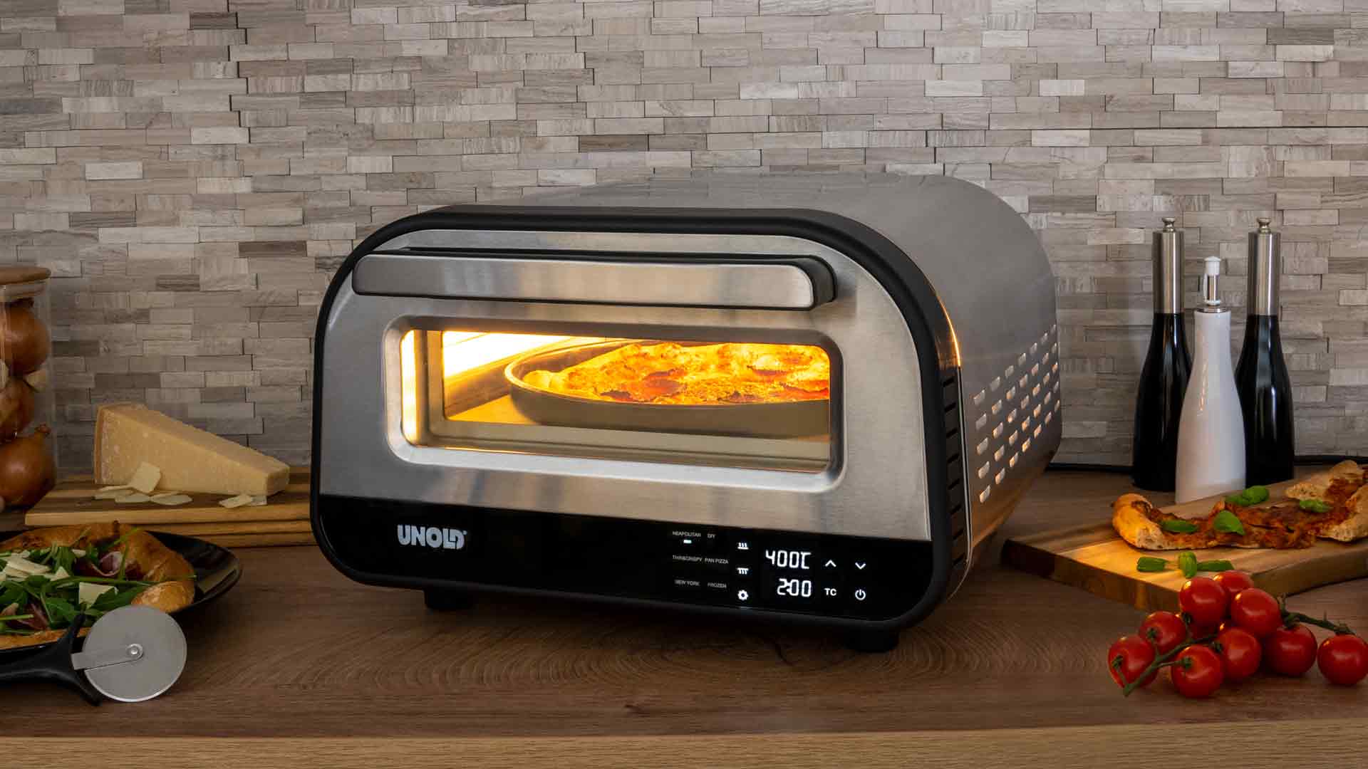 Ein kleiner Pizzaofen aus Edelstahl mit Digitalanzeige backt eine Pizza. Frische Pizza, Tomaten, Käse und Küchenutensilien liegen auf einer Holzarbeitsplatte in der Nähe. Im Hintergrund ist eine geflieste Küchenwand zu sehen.