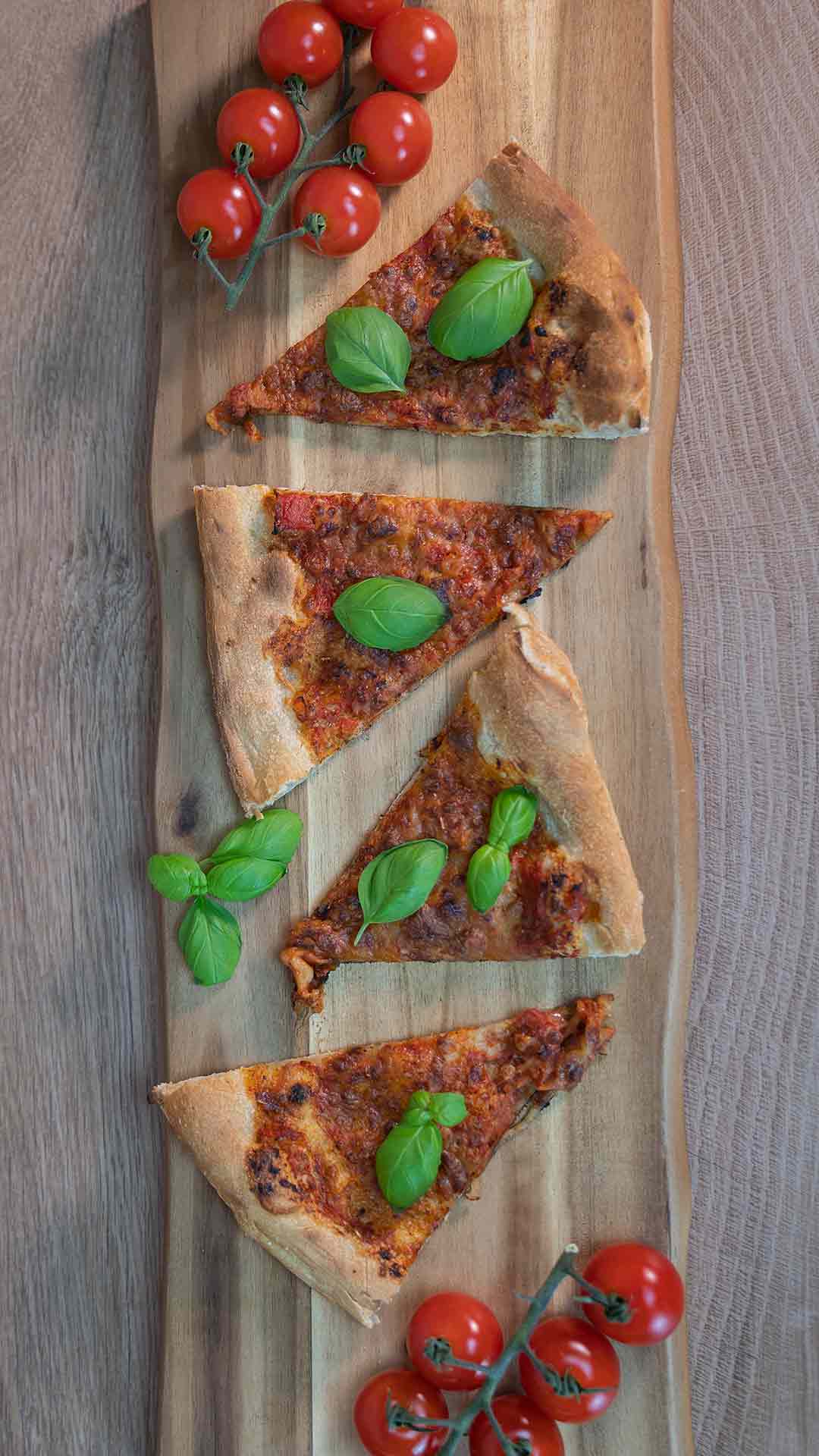 Vier Pizzastücke mit frischem Basilikum liegen auf einem hölzernen Servierbrett. An beiden Enden des Bretts befinden sich Trauben von Strauchtomaten. Der Hintergrund ist eine Holzoberfläche.