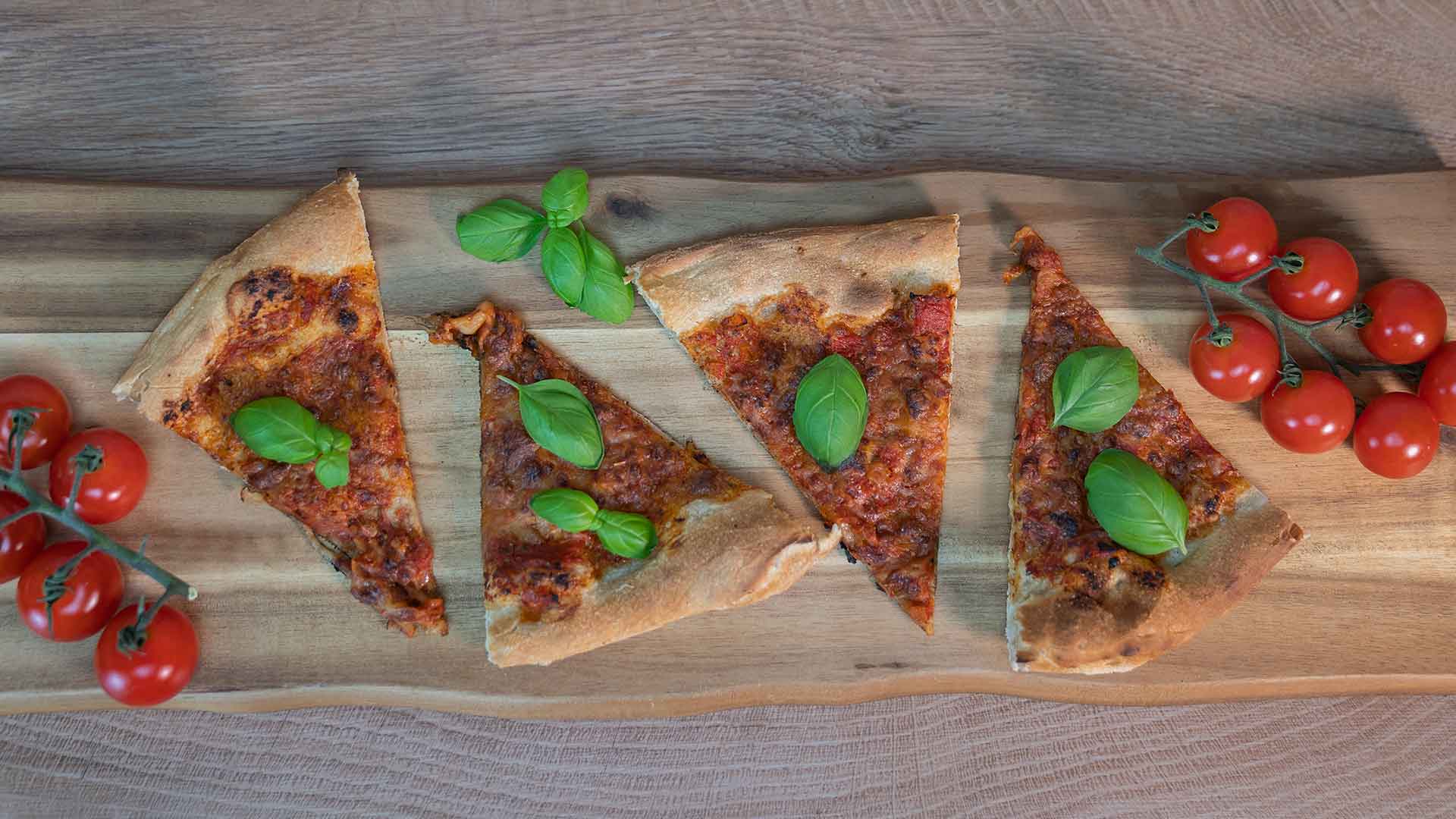 Fünf Pizzastücke mit Basilikumblättern darauf, angeordnet auf einem Holzbrett, mit Trauben frischer Kirschtomaten auf beiden Seiten des Bretts.