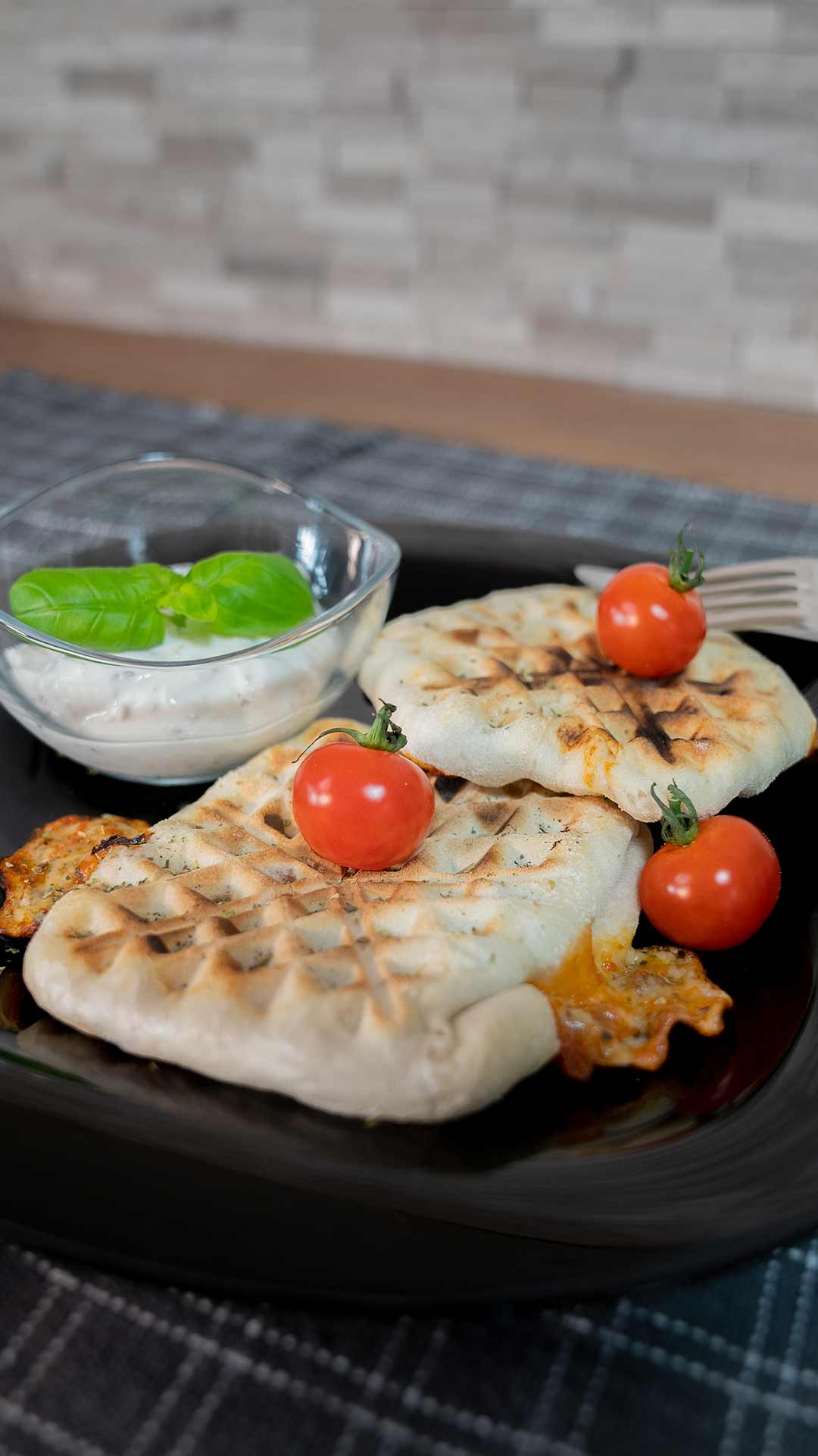 Zwei gegrillte Fladenbrote gefüllt mit geschmolzenem Käse und garniert mit frischen Kirschtomaten werden auf einem schwarzen Teller neben einer kleinen Schüssel cremigen Dip mit einem Basilikumblatt serviert.