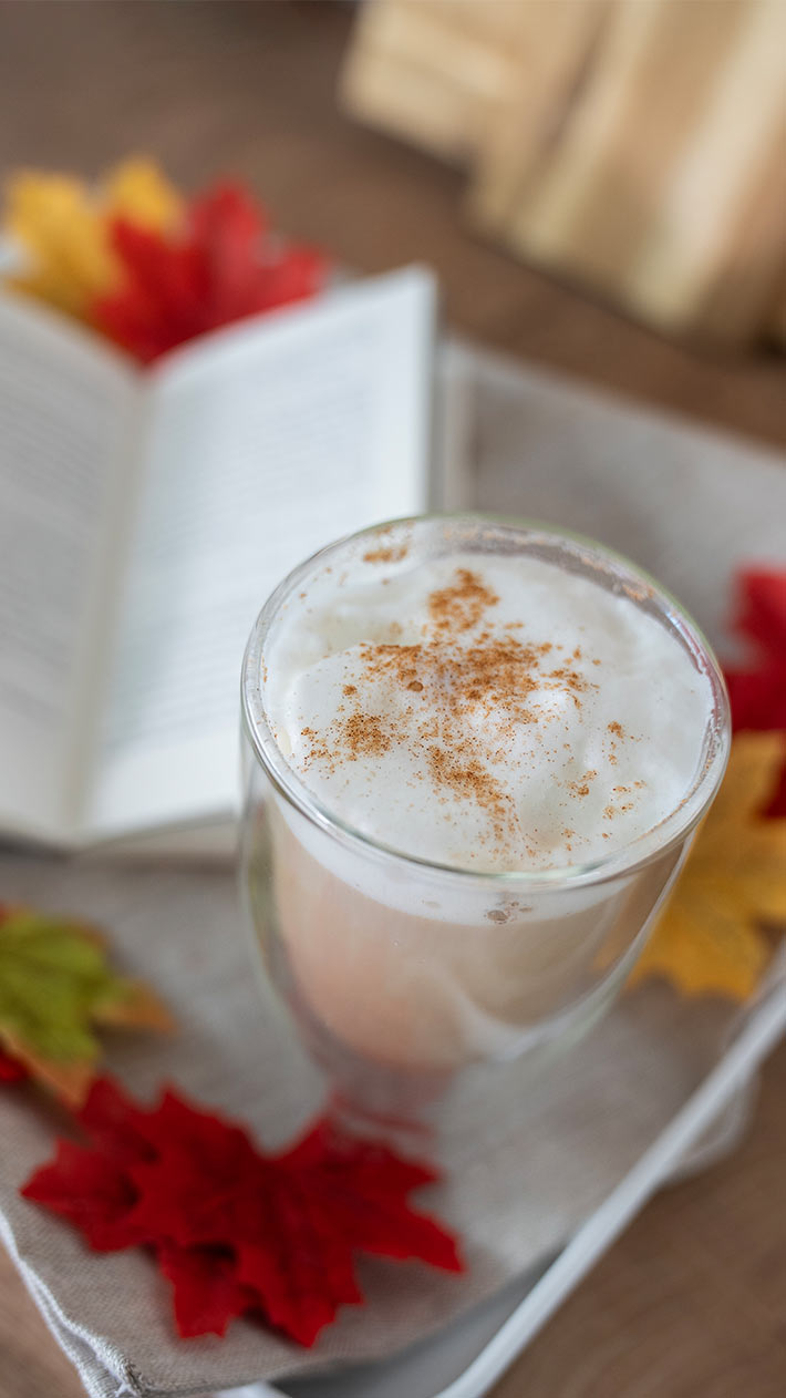Eine Glastasse gefüllt mit schaumigem Latte mit Zimt, auf einem Tablett mit Herbstblättern und einem offenen Buch im Hintergrund.