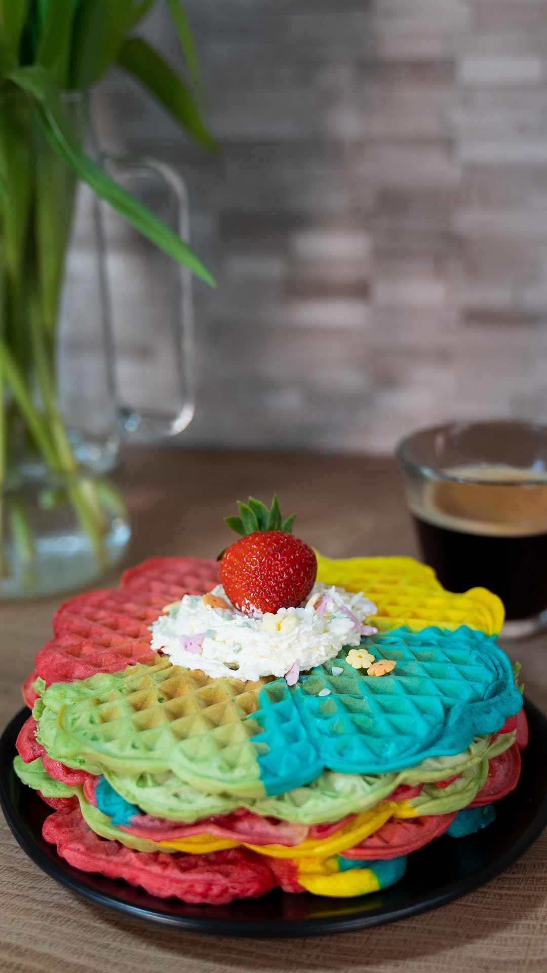 Ein Stapel bunter Regenbogenwaffeln mit Schlagsahne und einer ganzen Erdbeere liegt auf einem schwarzen Teller, im Hintergrund stehen eine Tasse Kaffee und grüne Lilien.