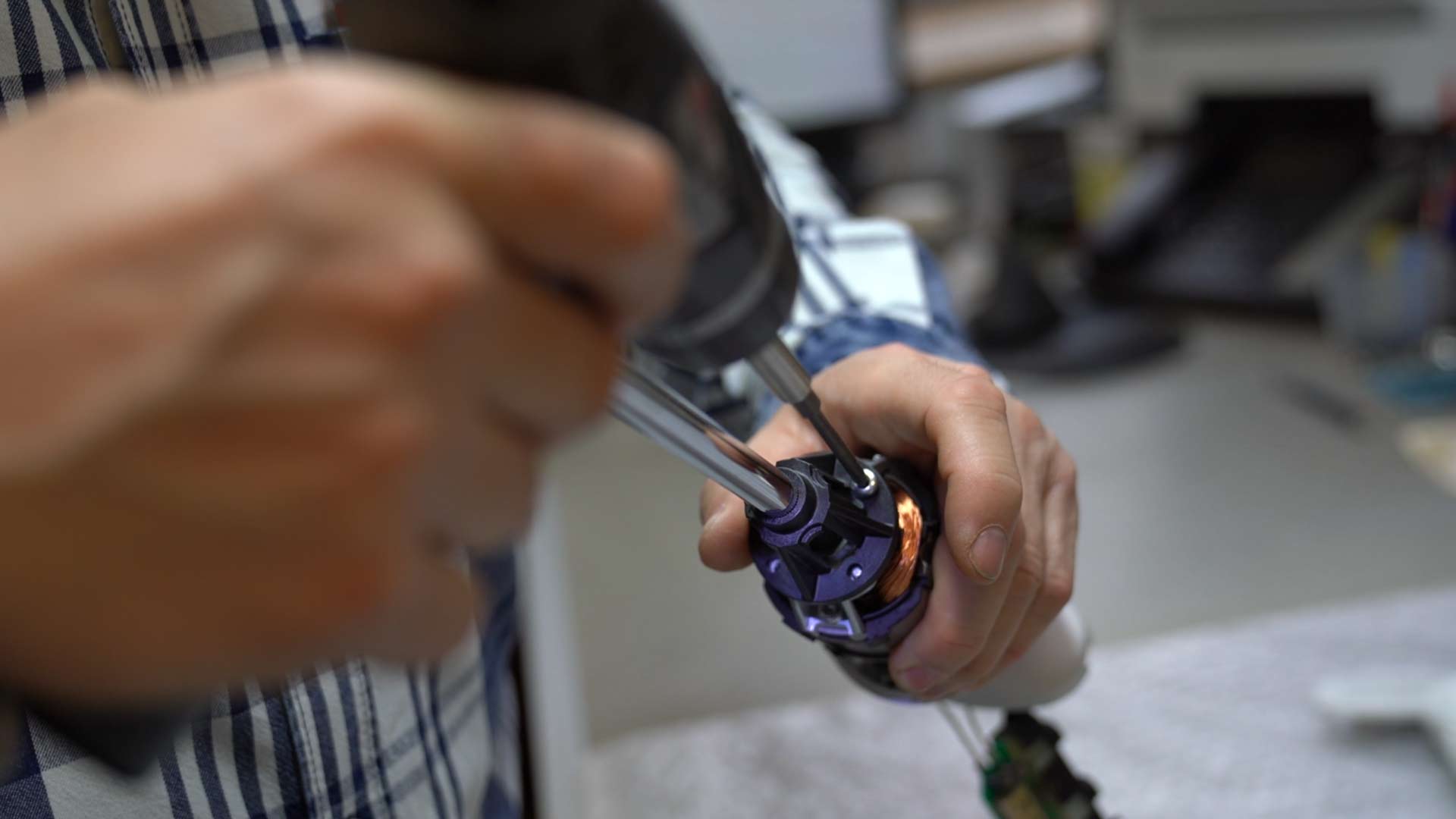 Nahaufnahme einer Person, die mit einer Bohrmaschine ein kleines mechanisches Gerät zusammenbaut oder repariert. Die Hände sind scharf dargestellt, der Hintergrund ist unscharf und zeigt einen Arbeitsbereich in einem Innenbereich.