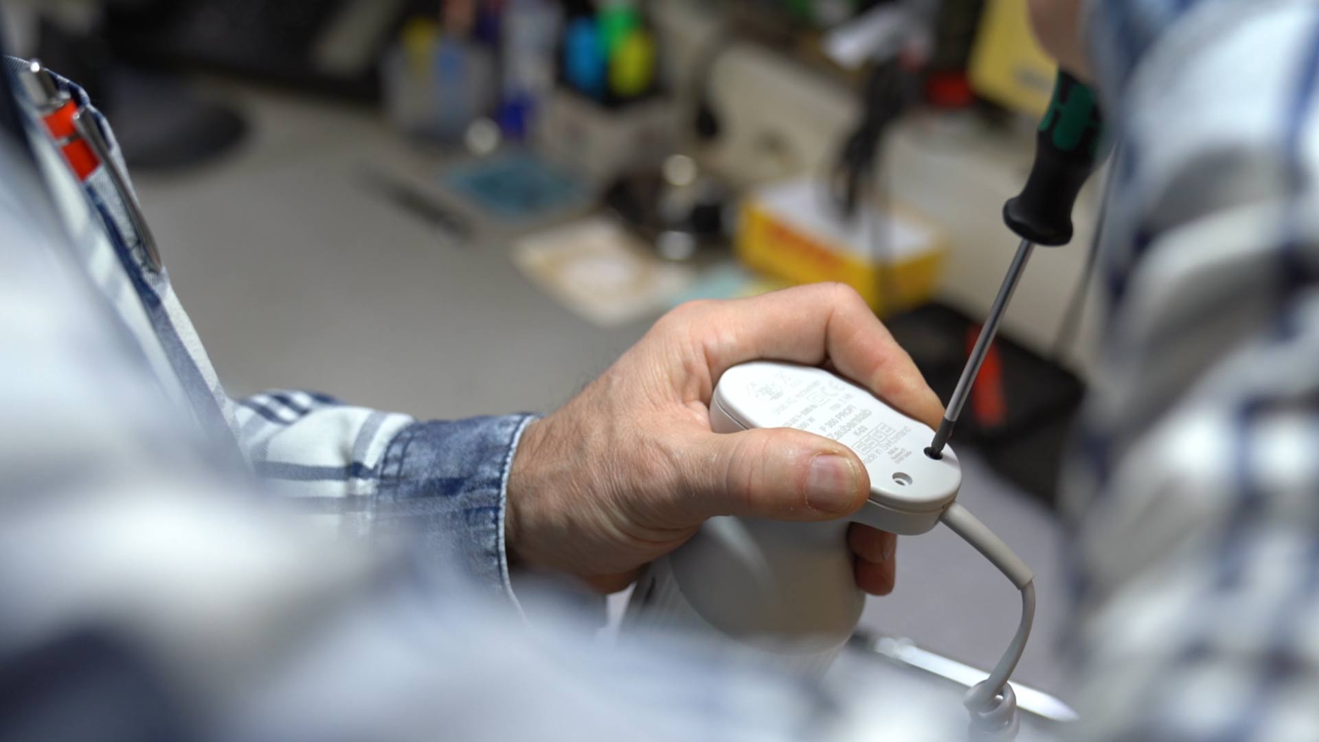 Eine Person öffnet mit einem Schraubenzieher die Unterseite einer kabelgebundenen Computermaus. Im Hintergrund sind verschiedene Werkzeuge und Objekte unscharf dargestellt.