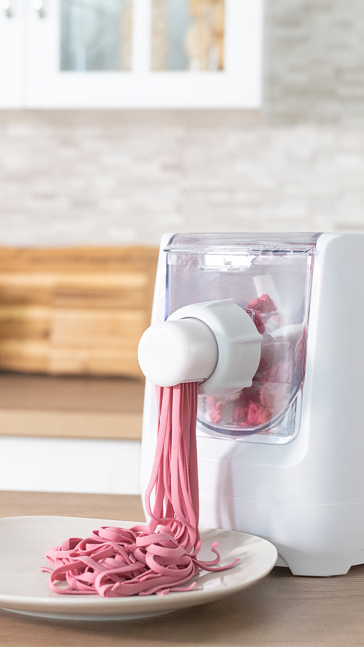 Eine weiße Nudelmaschine presst hellrosa Nudeln auf einen Teller auf einer Küchentheke, im Hintergrund sind ein verschwommenes Holzschneidebrett und Schränke zu sehen.