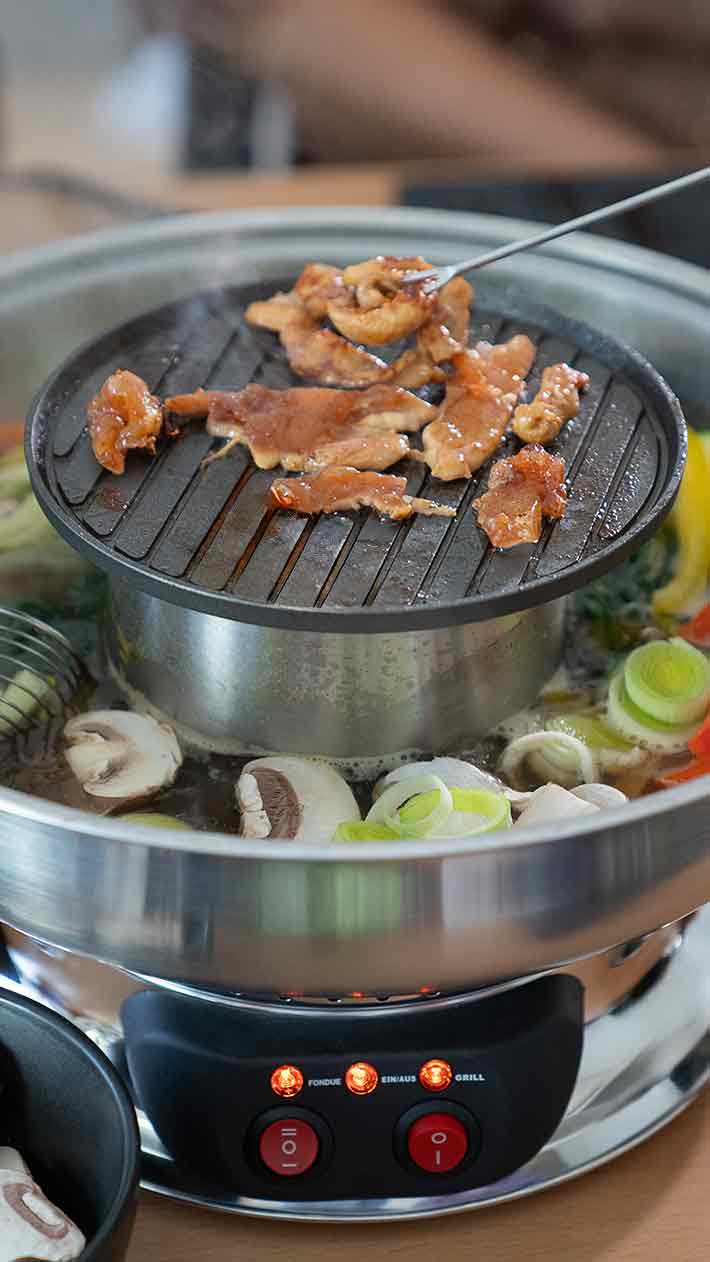 Nahaufnahme eines Tischgrills mit aufgeschnittenem Fleisch, das obenauf gart, und verschiedenes Gemüse, darunter Pilze und Lauch, das in Brühe köchelt. Am Gerät leuchten rote Kontrollleuchten.