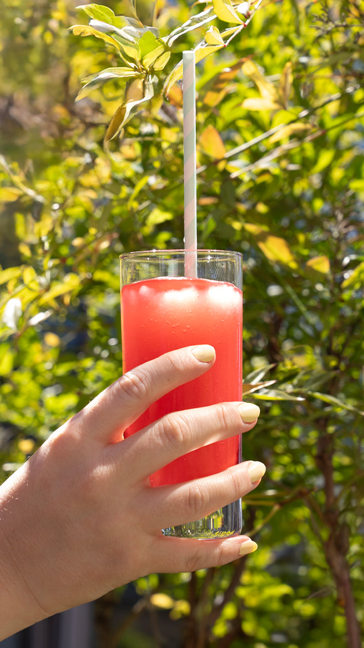 Eine Hand mit blassgelbem Nagellack hält ein Glas mit leuchtend rotem Slushy-Getränk mit einem gestreiften Strohhalm vor einem Hintergrund aus sonnenbeschienenen grünen Blättern.