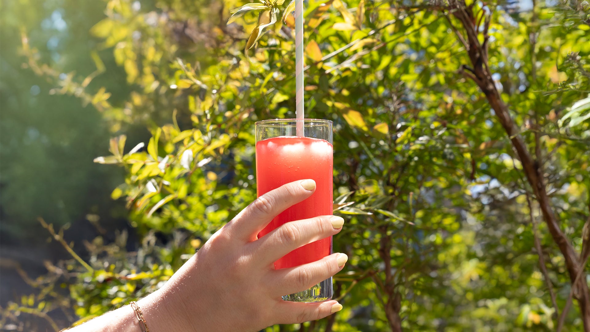Eine Hand, die ein Glas mit einem leuchtend roten, eisgekühlten Getränk mit einem Strohhalm hält, steht im Freien im Sonnenlicht mit grünen, belaubten Bäumen im Hintergrund.