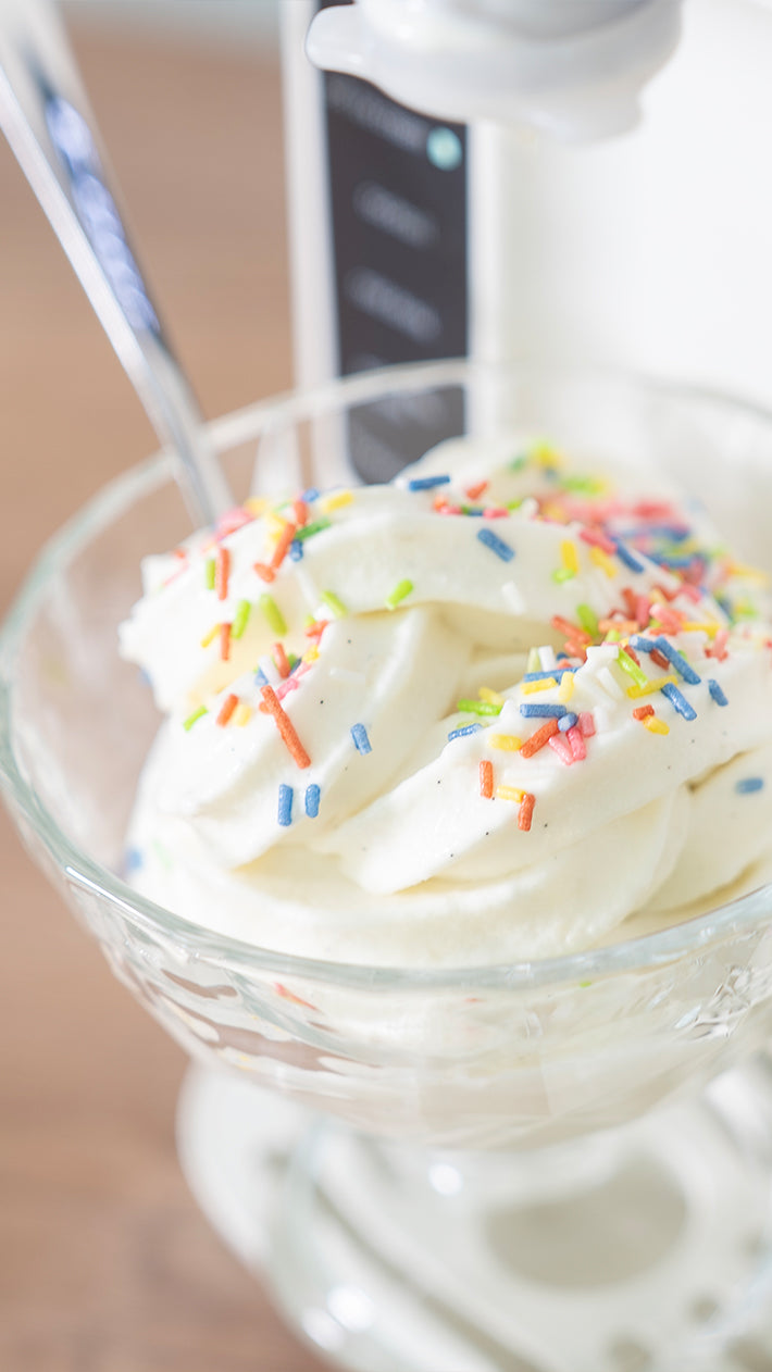 Eine Glasschüssel, gefüllt mit gewirbeltem Vanille-Softeis, bestreut mit bunten Regenbogenstreuseln, mit einem Metalllöffel an der Seite.