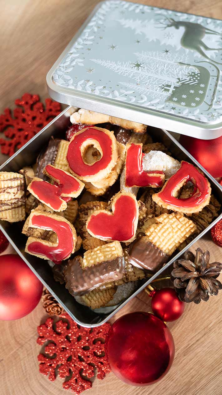 Eine offene Dose mit verschiedenen Weihnachtsplätzchen, einige davon mit roten Zuckergussbuchstaben „HOLID“ und einem Herz verziert. Die Dose ist von roten Ornamenten, Tannenzapfen und festlicher Dekoration umgeben.