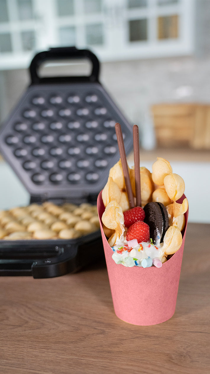Eine Bubble-Waffel in einem rosa Becher, garniert mit Schlagsahne, Himbeeren, einem Oreo-Keks, Schokoriegeln und bunten Süßigkeiten, steht auf einem Holztisch mit einem Waffeleisen und Küchenhintergrund.