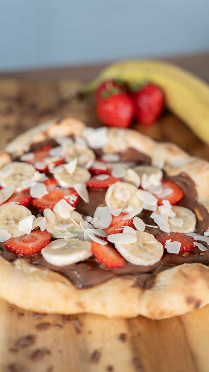 Eine Dessertpizza mit Schokoladenaufstrich, Bananenscheiben, Erdbeerscheiben und Mandelblättchen liegt auf einem Holzbrett, im Hintergrund sind ganze Erdbeeren und eine Banane zu sehen.
