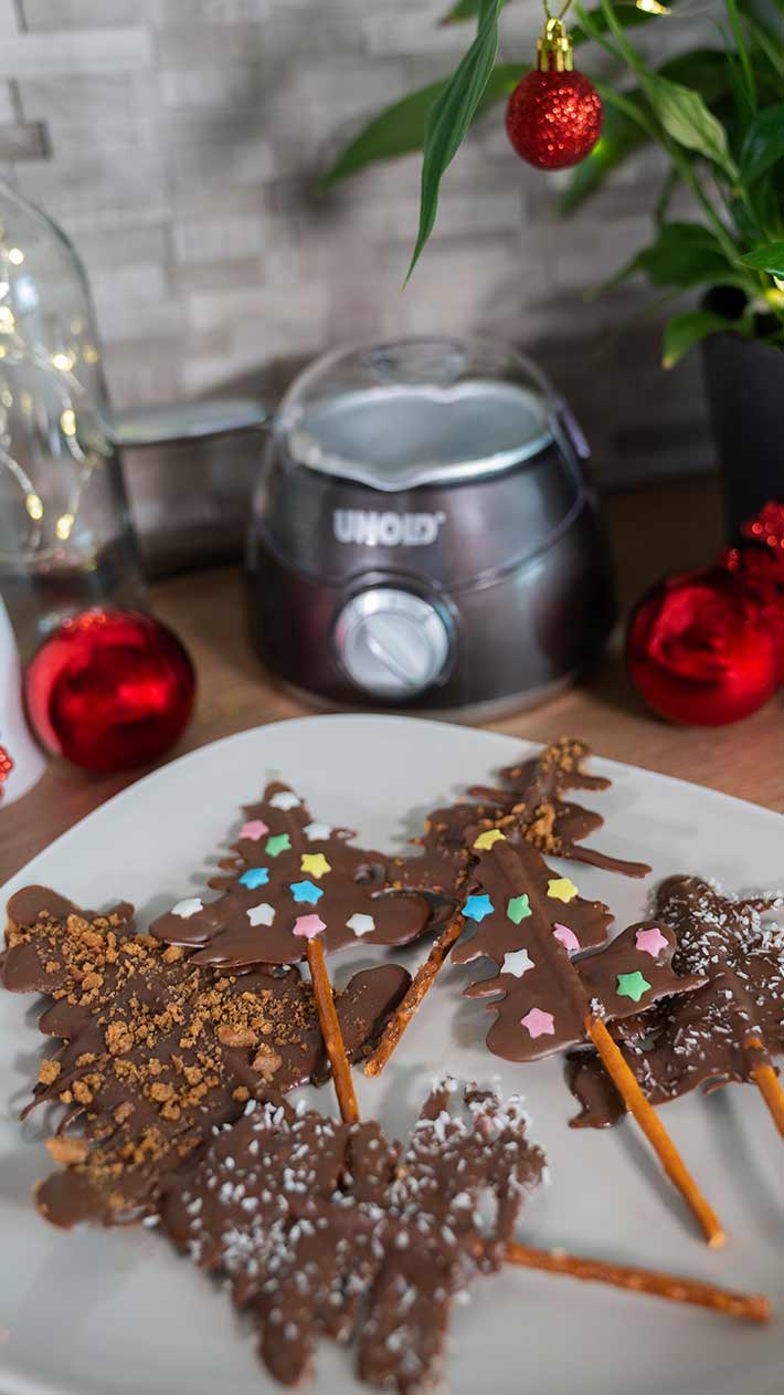 Ein Teller mit Schokoladen-Weihnachtsbaumleckereien aus Salzstangen, dekoriert mit bunten Streuseln und Toppings, steht auf einem Tisch mit festlichem Schmuck und einer Schokoladenschmelzmaschine im Hintergrund.