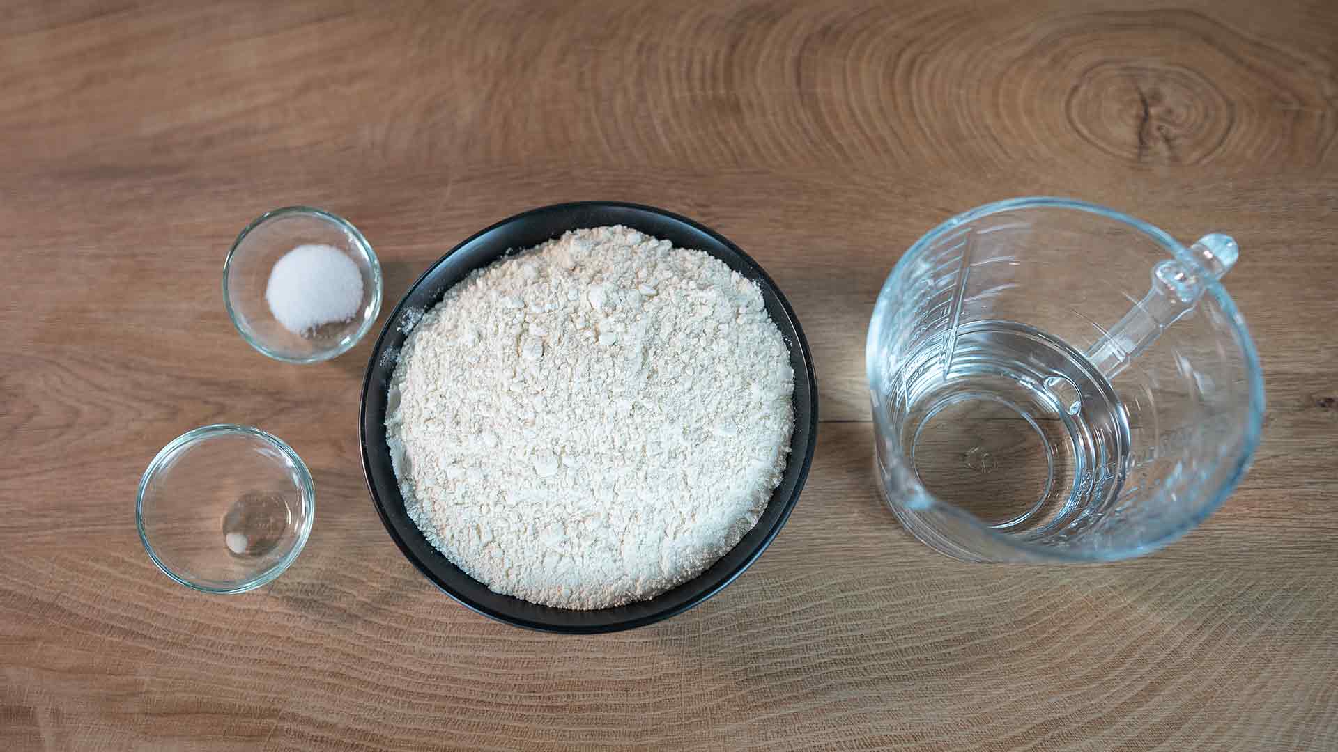Auf einer Holzfläche stehen eine Schüssel Mehl, ein Glaskrug Wasser und zwei kleine Glasschüsseln mit Salz und Hefe.