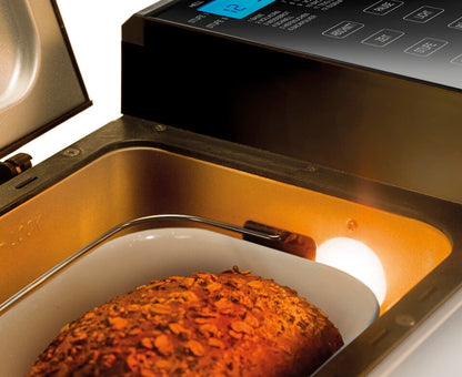Eine Nahaufnahme des Unold Backmeister® Top Edition Brotbackautomaten mit geöffnetem Deckel zeigt ein im Inneren backendes Körnerbrot. Das digitale Bedienfeld und die Tasten befinden sich auf der rechten Seite des Geräts.