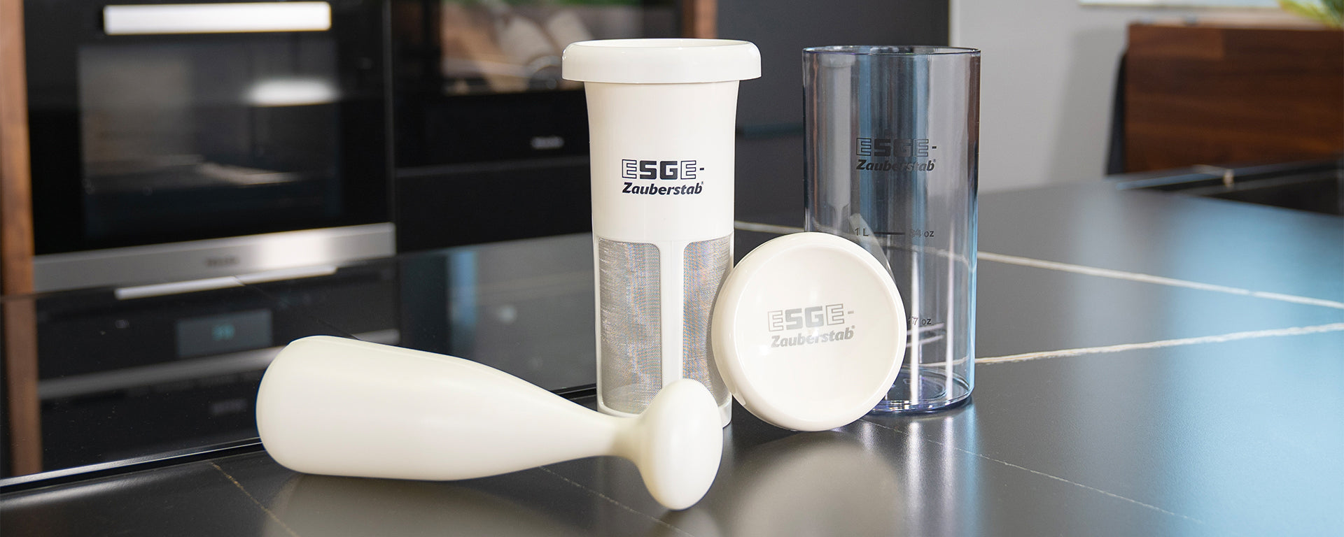 Auf einer Küchenarbeitsplatte sind Teile eines Küchengeräts zu sehen, darunter ein weißer Kolben, ein transparenter Behälter, ein weißer Deckel und ein zylindrischer Filter mit dem Schriftzug „EGE Zauberstab“. Im Hintergrund ist eine moderne Küche zu sehen.
