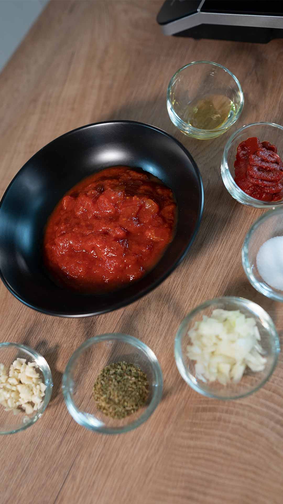 Eine schwarze Schüssel mit Tomatensoße ist von kleinen Glasschalen umgeben, die gehackte Zwiebeln, gehackten Knoblauch, getrocknete Kräuter, Tomatenmark, Olivenöl und Salz enthalten, alles auf einer Holzoberfläche angeordnet.