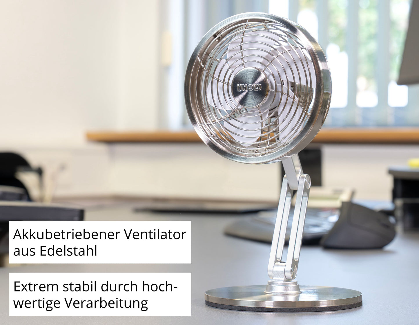 Der Unold AG Akku-Tischventilator Steel Flex, ein kompakter, akkubetriebener Edelstahlventilator, steht auf einem Bürotisch. Deutsche Etiketten weisen auf den wiederaufladbaren Akku und die hochwertige Verarbeitung hin. Unscharfe Monitore und Tastaturen erscheinen im Hintergrund.