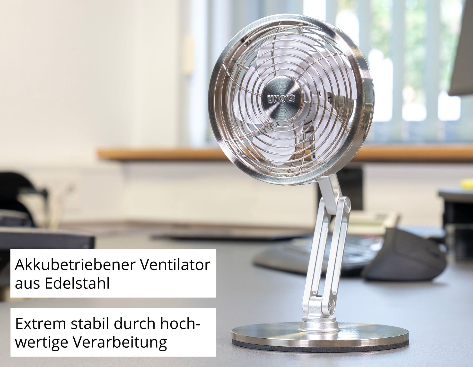 Der Unold AG Akku-Tischventilator Steel Flex, ein kompakter, akkubetriebener Edelstahlventilator, steht auf einem Bürotisch. Deutsche Etiketten weisen auf den wiederaufladbaren Akku und die hochwertige Verarbeitung hin. Unscharfe Monitore und Tastaturen erscheinen im Hintergrund.