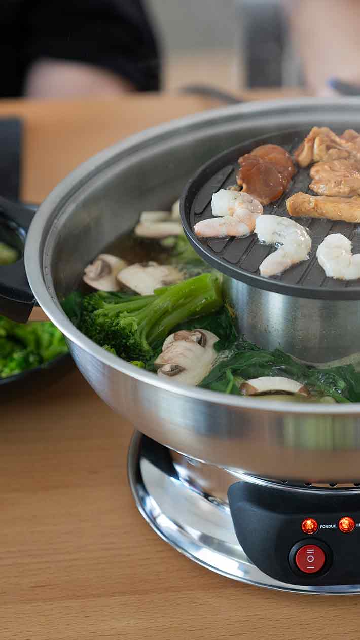 Auf einem Tisch steht ein Eintopf, in dem Gemüse und Pilze in Brühe kochen, während darüber auf einem Grill Garnelen und Fleischstücke garen.