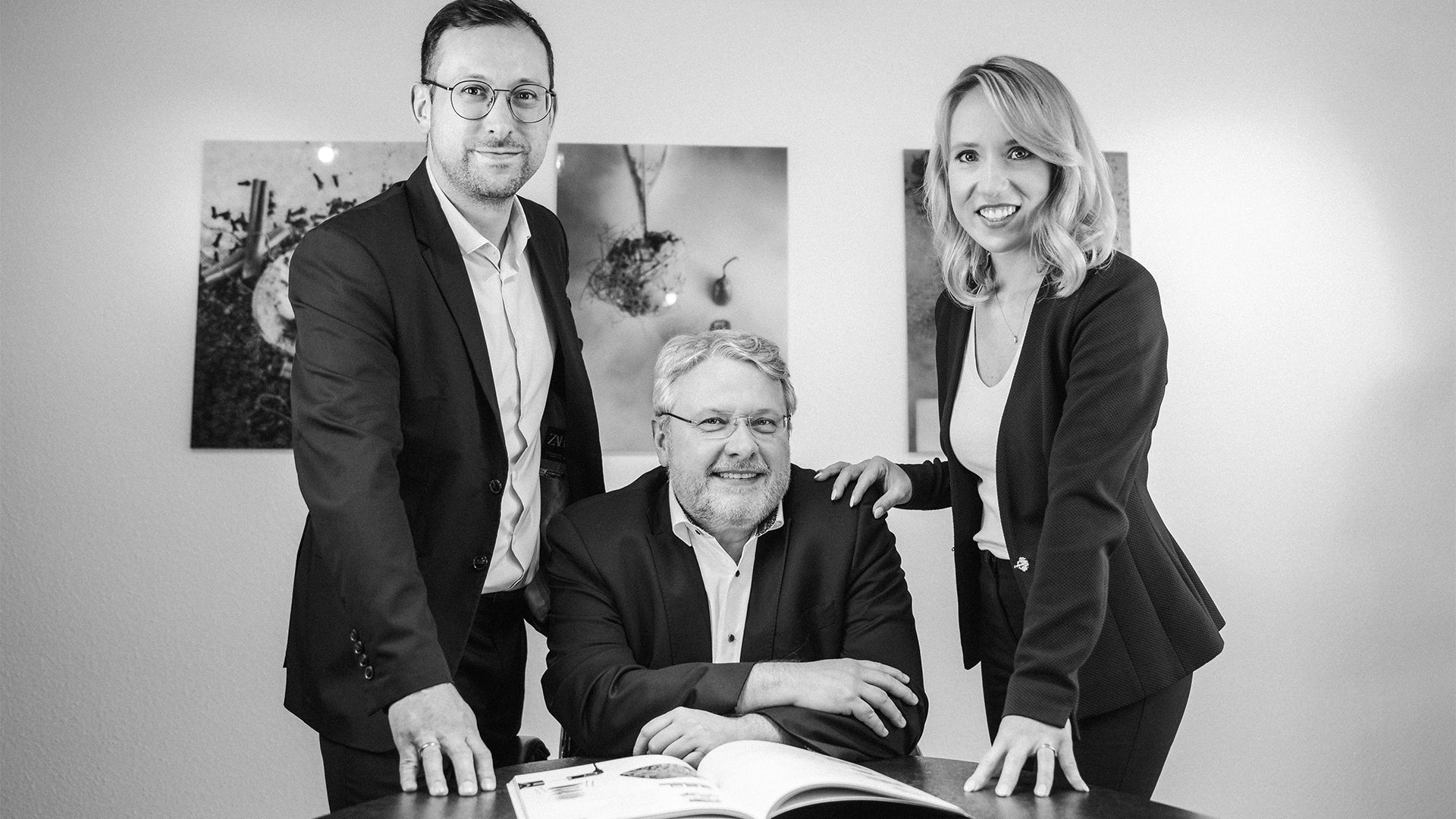 Drei Personen in Geschäftskleidung, zwei stehend und eine sitzend an einem Tisch mit einem offenen Buch, posieren und lächeln für die Kamera in einem modernen Büroumfeld mit gerahmter Wandkunst im Hintergrund.