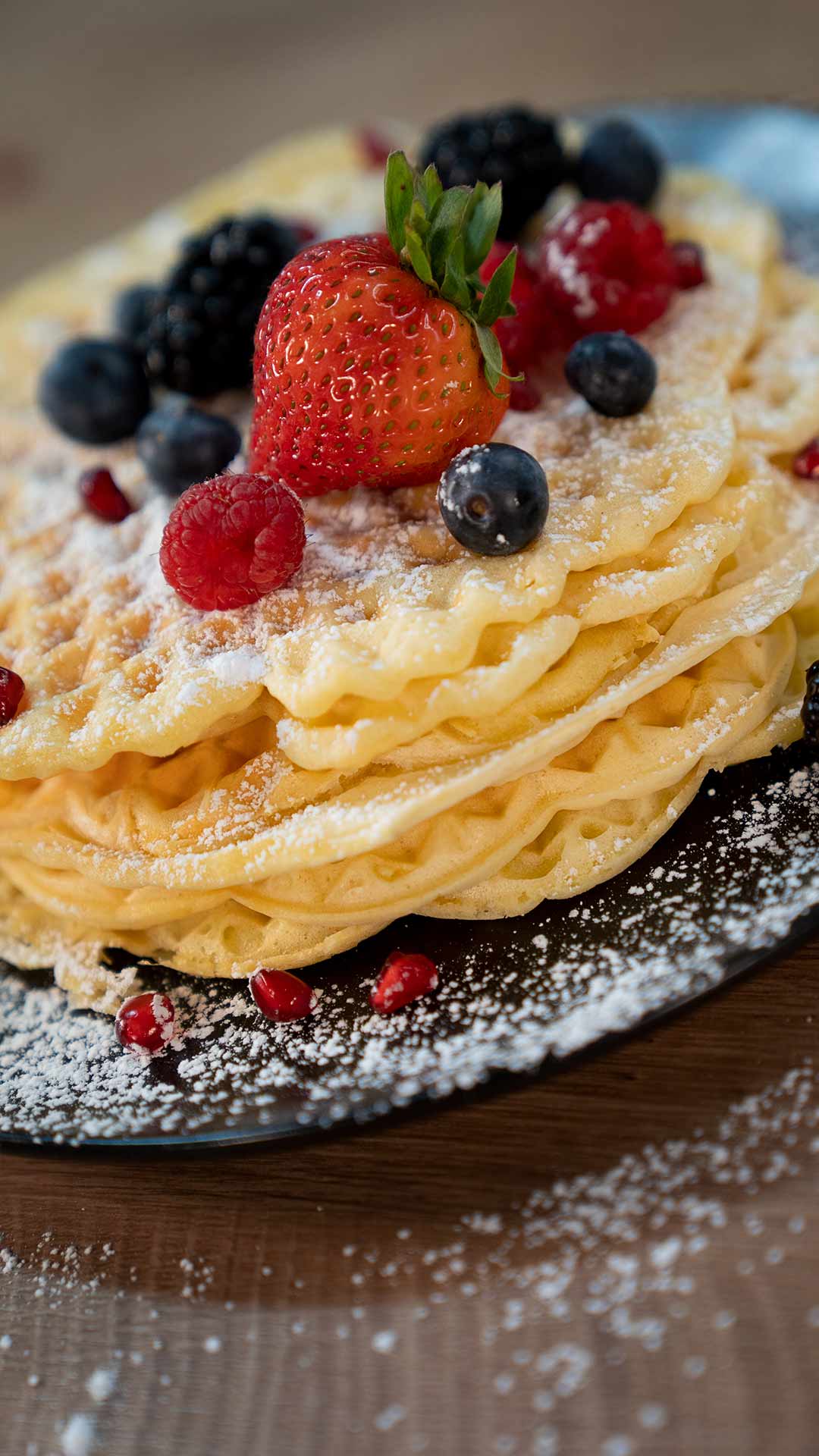 Ein Stapel Waffeln mit Erdbeeren, Blaubeeren, Himbeeren, Brombeeren und Puderzucker auf einem dunklen Teller, darüber verstreut einige Beeren und Puderzucker.