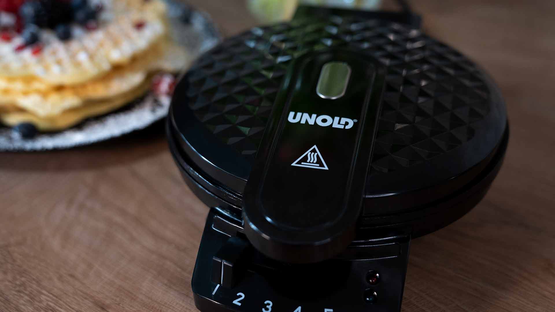 Auf einer Holzoberfläche steht ein schwarzes Waffeleisen von UNOLD, im Hintergrund ist ein Teller mit goldenen Waffeln und Beeren zu sehen.