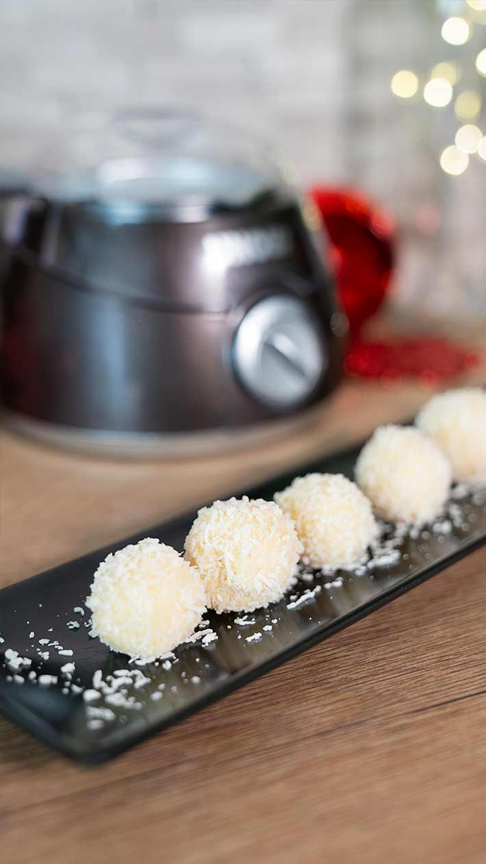 Fünf weiße Dessertkugeln mit Kokosnussüberzug liegen in einer Reihe auf einem schwarzen rechteckigen Teller, darüber gestreut mit Kokosraspeln. Im Hintergrund sind ein verschwommenes Küchengerät und festliche Lichter zu sehen.