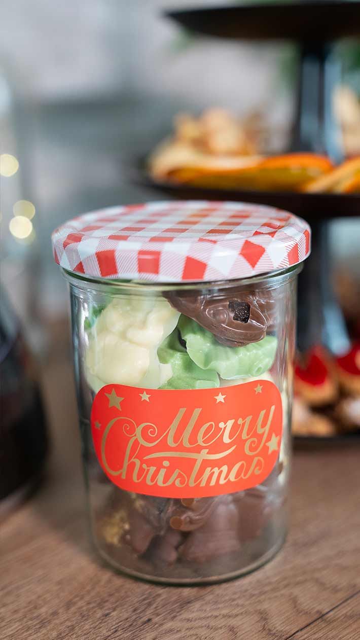 Ein Glas mit rot-weiß kariertem Deckel, gefüllt mit verschiedenen Pralinen und Süßigkeiten, trägt auf der Vorderseite ein festliches „Frohe Weihnachten“-Etikett. Das Glas steht auf einer Holzoberfläche, im Hintergrund sind unscharfe Leckereien zu sehen.